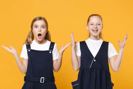 Two young little girls 12-13 years old in white t-shirt blue school uniform dresses isolated on yellow background children studio portrait Childhood kids education lifestyle concept Mock up copy spaceの写真素材