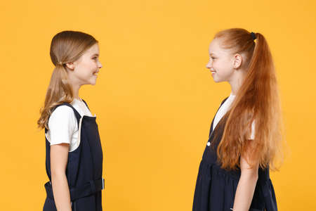 Two young little girls 12-13 years old in white t-shirt blue school uniform dresses isolated on yellow background children studio portrait Childhood kids education lifestyle concept Mock up copy spaceの写真素材
