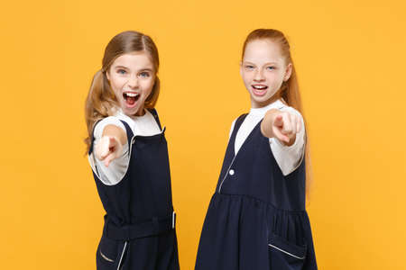 Two young little girls 12-13 years old in white t-shirt blue school uniform dresses isolated on yellow background children studio portrait Childhood kids education lifestyle concept Mock up copy spaceの写真素材