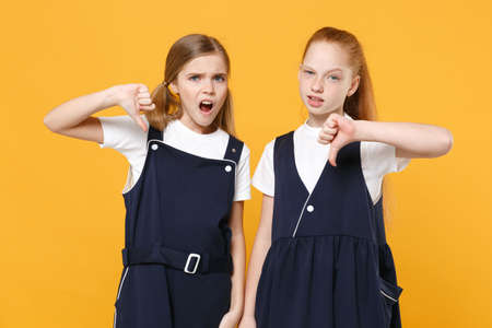 Two young little girls 12-13 years old in white t-shirt blue school uniform dresses isolated on yellow background children studio portrait Childhood kids education lifestyle concept Mock up copy spaceの写真素材