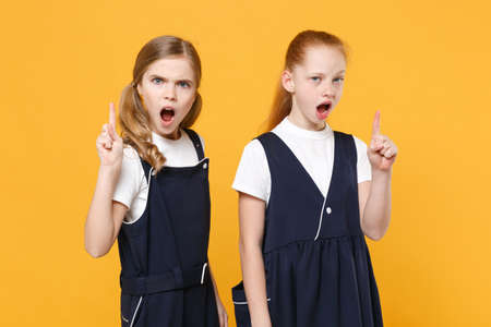 Two young little girls 12-13 years old in white t-shirt blue school uniform dresses isolated on yellow background children studio portrait Childhood kids education lifestyle concept Mock up copy spaceの写真素材