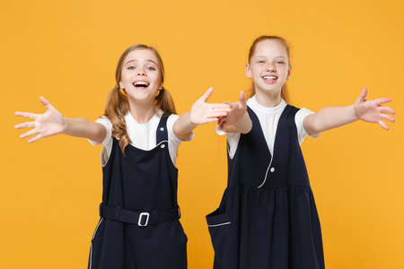 Two young little girls 12-13 years old in white t-shirt blue school uniform dresses isolated on yellow background children studio portrait Childhood kids education lifestyle concept Mock up copy spaceの写真素材