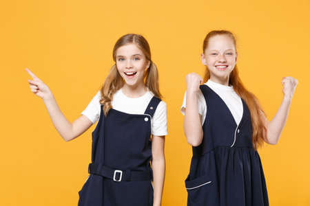 Two young little girls 12-13 years old in white t-shirt blue school uniform dresses isolated on yellow background children studio portrait Childhood kids education lifestyle concept Mock up copy spaceの写真素材