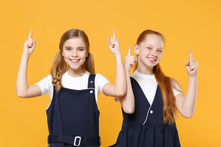 Two young little girls 12-13 years old in white t-shirt blue school uniform dresses isolated on yellow background children studio portrait Childhood kids education lifestyle concept Mock up copy spaceの写真素材