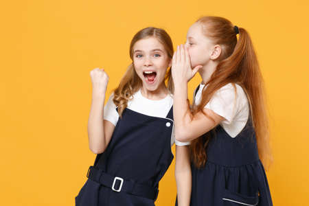 Two young little girls 12-13 years old in white t-shirt blue school uniform dresses isolated on yellow background children studio portrait Childhood kids education lifestyle concept Mock up copy spaceの写真素材
