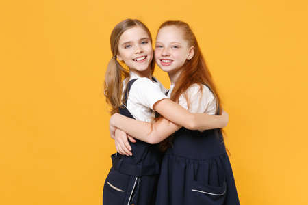 Two young little girls 12-13 years old in white t-shirt blue school uniform dresses isolated on yellow background children studio portrait Childhood kids education lifestyle concept Mock up copy spaceの写真素材