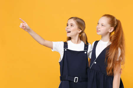 Two young little girls 12-13 years old in white t-shirt blue school uniform dresses isolated on yellow background children studio portrait Childhood kids education lifestyle concept Mock up copy spaceの写真素材