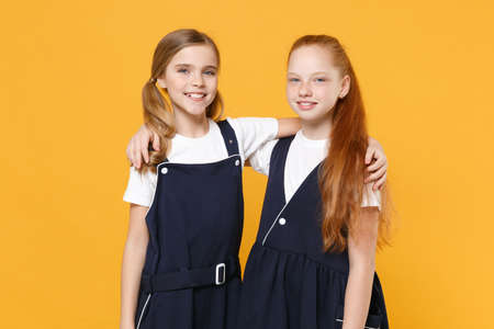 Two young little girls 12-13 years old in white t-shirt blue school uniform dresses isolated on yellow background children studio portrait Childhood kids education lifestyle concept Mock up copy spaceの写真素材