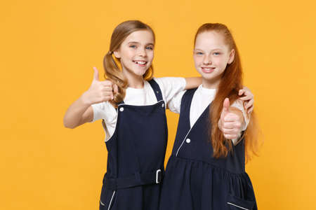 Two young little girls 12-13 years old in white t-shirt blue school uniform dresses isolated on yellow background children studio portrait Childhood kids education lifestyle concept Mock up copy spaceの写真素材