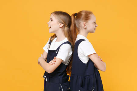 Two young little girls 12-13 years old in white t-shirt blue school uniform dresses isolated on yellow background children studio portrait Childhood kids education lifestyle concept Mock up copy spaceの写真素材
