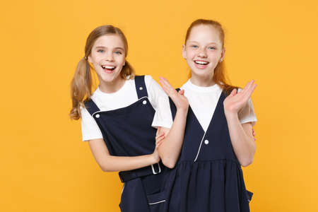 Two young little girls 12-13 years old in white t-shirt blue school uniform dresses isolated on yellow background children studio portrait Childhood kids education lifestyle concept Mock up copy spaceの写真素材