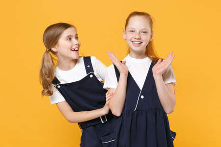Two young little girls 12-13 years old in white t-shirt blue school uniform dresses isolated on yellow background children studio portrait Childhood kids education lifestyle concept Mock up copy spaceの写真素材