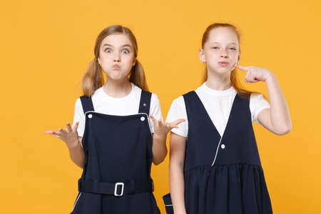 Two young little girls 12-13 years old in white t-shirt blue school uniform dresses isolated on yellow background children studio portrait Childhood kids education lifestyle concept Mock up copy spaceの写真素材
