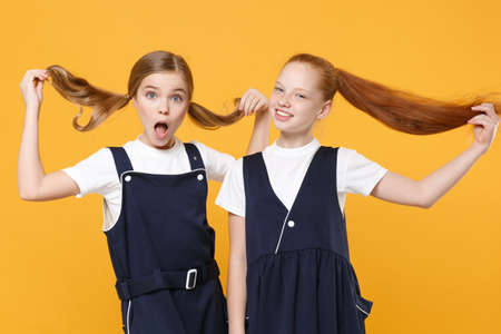 Two young little girls 12-13 years old in white t-shirt blue school uniform dresses isolated on yellow background children studio portrait Childhood kids education lifestyle concept Mock up copy spaceの写真素材