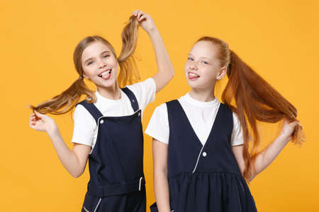 Two young little girls 12-13 years old in white t-shirt blue school uniform dresses isolated on yellow background children studio portrait Childhood kids education lifestyle concept Mock up copy spaceの写真素材