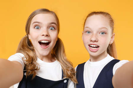 Two young little girls 12-13 years old in white t-shirt blue school uniform dresses isolated on yellow background children studio portrait Childhood kids education lifestyle concept Mock up copy spaceの写真素材