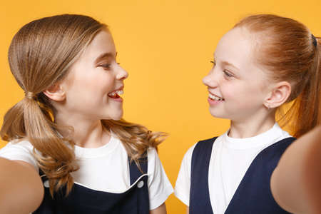 Two young little girls 12-13 years old in white t-shirt blue school uniform dresses isolated on yellow background children studio portrait Childhood kids education lifestyle concept Mock up copy spaceの写真素材
