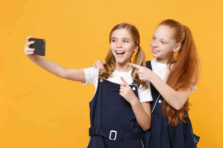 Two girls 12-13 years old in white t-shirt school uniform dresses doing selfie shot isolated on yellow background children studio portrait Childhood kids education lifestyle concept Mock up copy spaceの写真素材