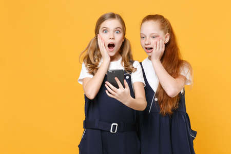 Two girls 12-13 years old in white t-shirt blue school dresses using mobile phone isolated on yellow background children studio portrait Childhood kids education lifestyle concept Mock up copy spaceの写真素材