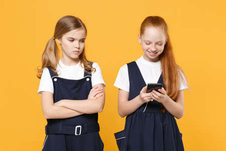 Two girls 12-13 years old in white t-shirt blue school dresses using mobile phone isolated on yellow background children studio portrait Childhood kids education lifestyle concept Mock up copy spaceの写真素材