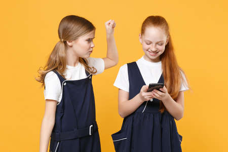 Two girls 12-13 years old in white t-shirt blue school dresses using mobile phone isolated on yellow background children studio portrait Childhood kids education lifestyle concept Mock up copy spaceの写真素材
