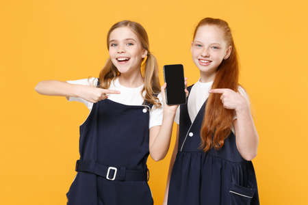 Two girls 12-13 years old in white t-shirt school uniform dresses hold mobile phone isolated on yellow background children studio portrait Childhood kids education lifestyle concept Mock up copy spaceの写真素材