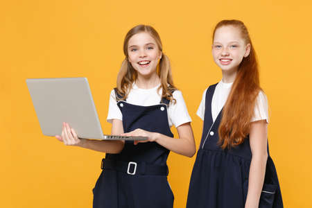 Two girls 12-13 years old in white t-shirt blue school uniform hold laptop pc computer isolated on yellow background children studio portrait Childhood education lifestyle concept Mock up copy spaceの写真素材