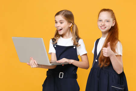 Two girls 12-13 years old in white t-shirt blue school uniform hold laptop pc computer isolated on yellow background children studio portrait Childhood education lifestyle concept Mock up copy spaceの写真素材