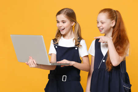 Two girls 12-13 years old in white t-shirt blue school uniform hold laptop pc computer isolated on yellow background children studio portrait Childhood education lifestyle concept Mock up copy spaceの写真素材