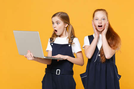 Two girls 12-13 years old in white t-shirt blue school uniform hold laptop pc computer isolated on yellow background children studio portrait Childhood education lifestyle concept Mock up copy spaceの写真素材