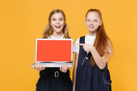 Two girls 12-13 years old in white t-shirt blue school uniform hold laptop pc computer isolated on yellow background children studio portrait Childhood education lifestyle concept Mock up copy spaceの写真素材