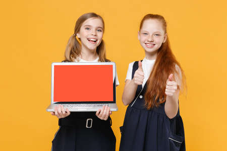 Two girls 12-13 years old in white t-shirt blue school uniform hold laptop pc computer isolated on yellow background children studio portrait Childhood education lifestyle concept Mock up copy spaceの写真素材