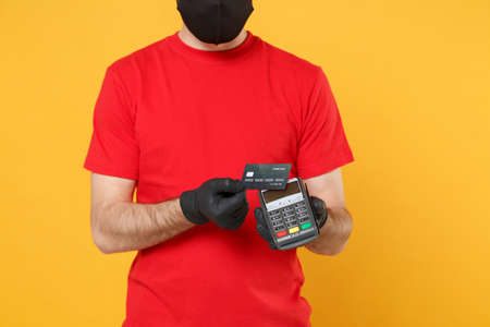 Delivery man in red cap blank t-shirt sterile mask gloves isolated on yellow background studio Guy employee hold bank payment terminal Service quarantine pandemic coronavirus virus 2019-ncov concept.の写真素材