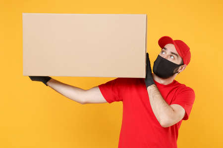 Delivery man employee in red cap blank t-shirt uniform face mask glove hold empty cardboard box isolated on yellow background studio Service quarantine pandemic coronavirus flu virus 2019-ncov conceptの写真素材