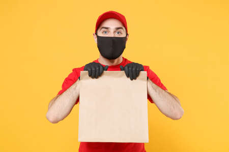 Delivery man employee in red cap blank t-shirt uniform mask glove hold craft paper packet with food isolated on yellow background studio Service quarantine pandemic coronavirus virus 2019-ncov conceptの写真素材