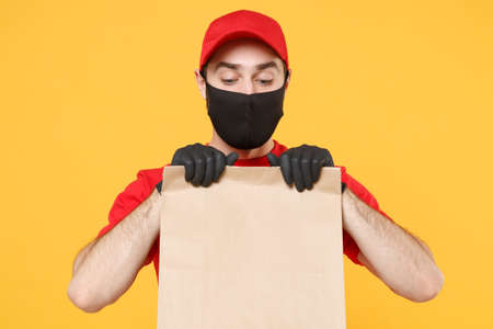 Delivery man employee in red cap blank t-shirt uniform mask glove hold craft paper packet with food isolated on yellow background studio Service quarantine pandemic coronavirus virus 2019-ncov conceptの写真素材