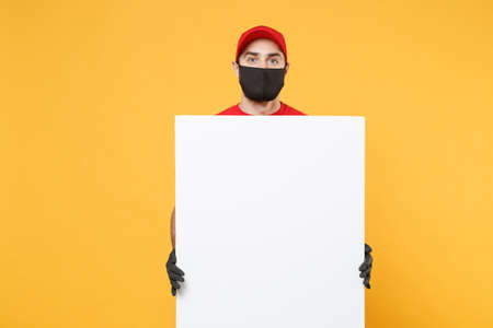 Delivery man in red cap t-shirt uniform sterile face mask gloves hold blank sign board isolated on yellow background studio Guy employee Service quarantine pandemic coronavirus virus 2019-ncov conceptの写真素材
