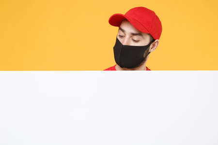 Delivery man in red cap t-shirt uniform sterile face mask gloves hold blank sign board isolated on yellow background studio Guy employee Service quarantine pandemic coronavirus virus 2019-ncov conceptの写真素材