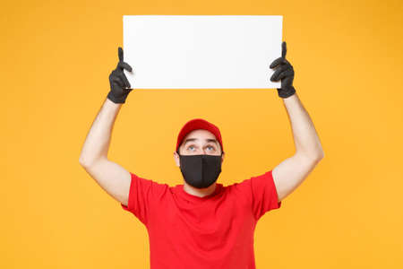 Delivery man in red cap t-shirt uniform sterile face mask gloves hold blank sign board isolated on yellow background studio Guy employee Service quarantine pandemic coronavirus virus 2019-ncov conceptの写真素材