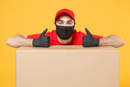 Delivery man employee in red cap blank t-shirt uniform face mask glove hold empty cardboard box isolated on yellow background studio Service quarantine pandemic coronavirus flu virus 2019-ncov conceptの写真素材