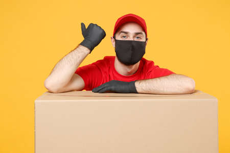 Delivery man employee in red cap blank t-shirt uniform face mask glove hold empty cardboard box isolated on yellow background studio Service quarantine pandemic coronavirus flu virus 2019-ncov conceptの写真素材