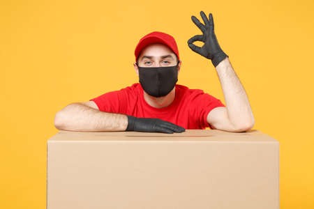 Delivery man employee in red cap blank t-shirt uniform face mask glove hold empty cardboard box isolated on yellow background studio Service quarantine pandemic coronavirus flu virus 2019-ncov conceptの写真素材