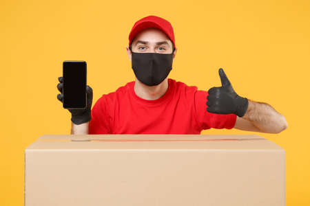 Delivery man employee in red cap blank t-shirt uniform face mask glove hold empty cardboard box isolated on yellow background studio Service quarantine pandemic coronavirus flu virus 2019-ncov conceptの写真素材