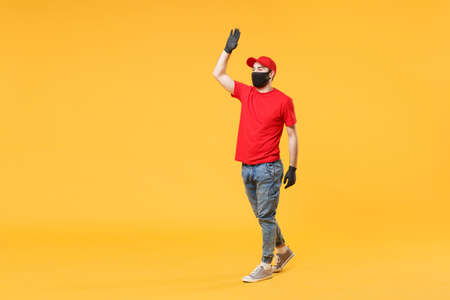 Full length delivery man in red cap t-shirt uniform sterile face mask gloves isolated on yellow background studio Guy employee courier Service quarantine pandemic coronavirus virus 2019-ncov concept.の写真素材