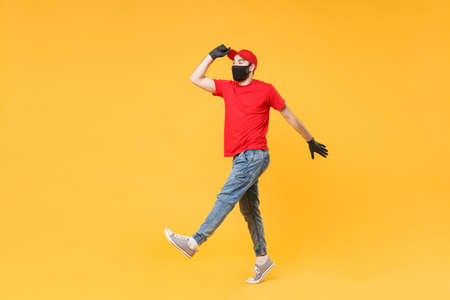Full length delivery man in red cap t-shirt uniform sterile face mask gloves isolated on yellow background studio Guy employee courier Service quarantine pandemic coronavirus virus 2019-ncov concept.の写真素材