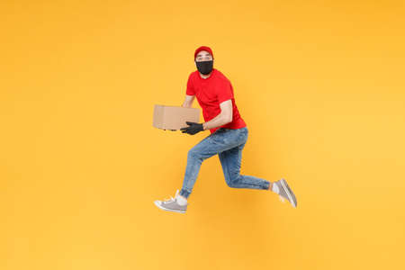 Fun jumping delivery man in red cap t-shirt uniform sterile face mask gloves isolated on yellow background studio Guy employee courier Service quarantine pandemic coronavirus virus 2019-ncov concept.の写真素材