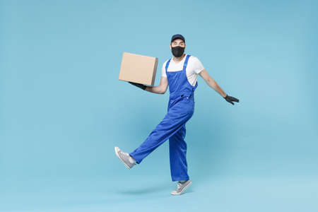 Full length delivery man in cap t-shirt uniform sterile face mask gloves isolated on blue background studio Guy employee courier hold box Service pandemic coronavirus virus covid-19 2019-ncov concept.の写真素材