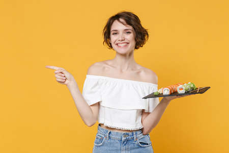 Young brunette woman girl in casual clothes hold in hand makizushi sushi roll served on black plate traditional japanese food isolated on yellow background studio portrait. People lifestyle concept.の写真素材