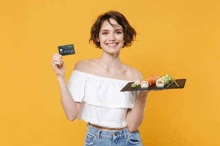 Young woman girl in casual clothes hold in hand credit card makizushi sushi roll served on black plate traditional japanese food isolated on yellow background studio portrait. People lifestyle conceptの写真素材