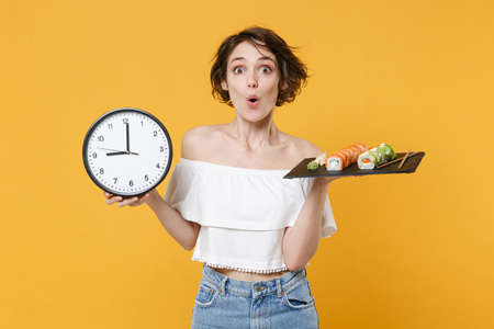 Young fun woman girl in casual clothes hold in hand clock makizushi sushi roll served on black plate traditional japanese food isolated on yellow background studio portrait. People lifestyle concept.の写真素材